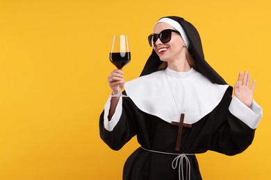 Happy woman in nun habit and sunglasses holding glass of wine against orange background. Space for text