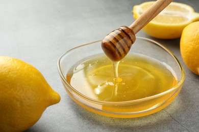 Photo of Dripping sweet honey from dipper into bowl and fresh lemons on grey table, closeup