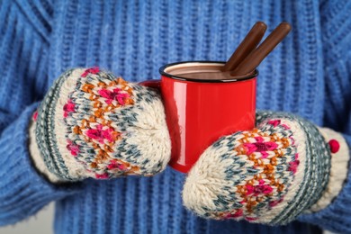 Photo of Woman in knitted mittens holding cup of delicious hot chocolate with cocoa sticks, closeup