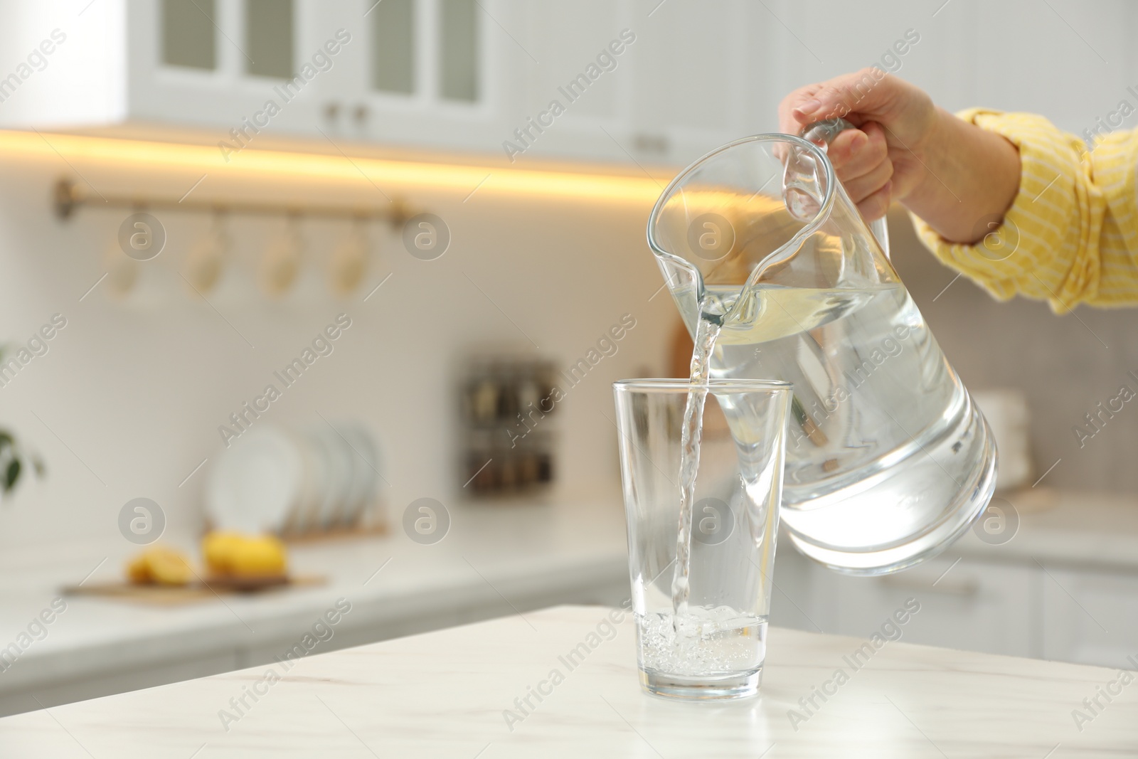 Photo of Woman pouring water from jug into glass at white table in kitchen, closeup. Space for text