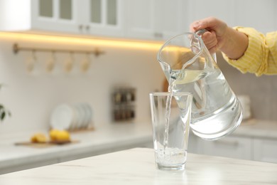 Photo of Woman pouring water from jug into glass at white table in kitchen, closeup. Space for text