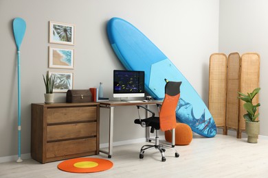 Photo of Stylish workplace with modern computer and SUP board near light wall in room. Interior design