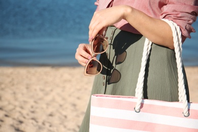 Woman holding bag and sunglasses on beach, closeup. Summer vacation