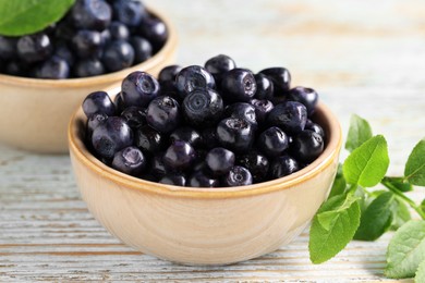 Photo of Bowls with tasty fresh bilberries and leaves on old light blue wooden table, closeup