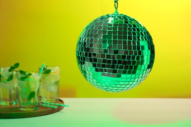 Shiny disco ball hanging over white table with cocktails against yellow background