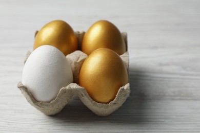 Carton with golden eggs and ordinary one on white wooden table, closeup. Space for text