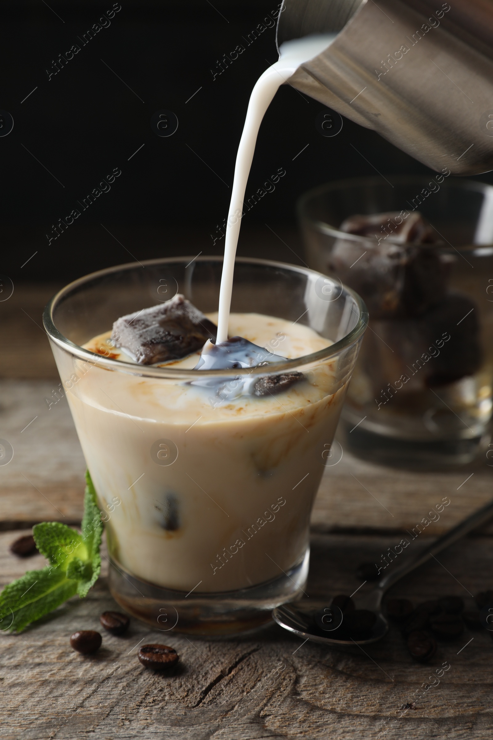 Photo of Pouring milk into glass of delicious iced coffee, mint and beans on wooden table