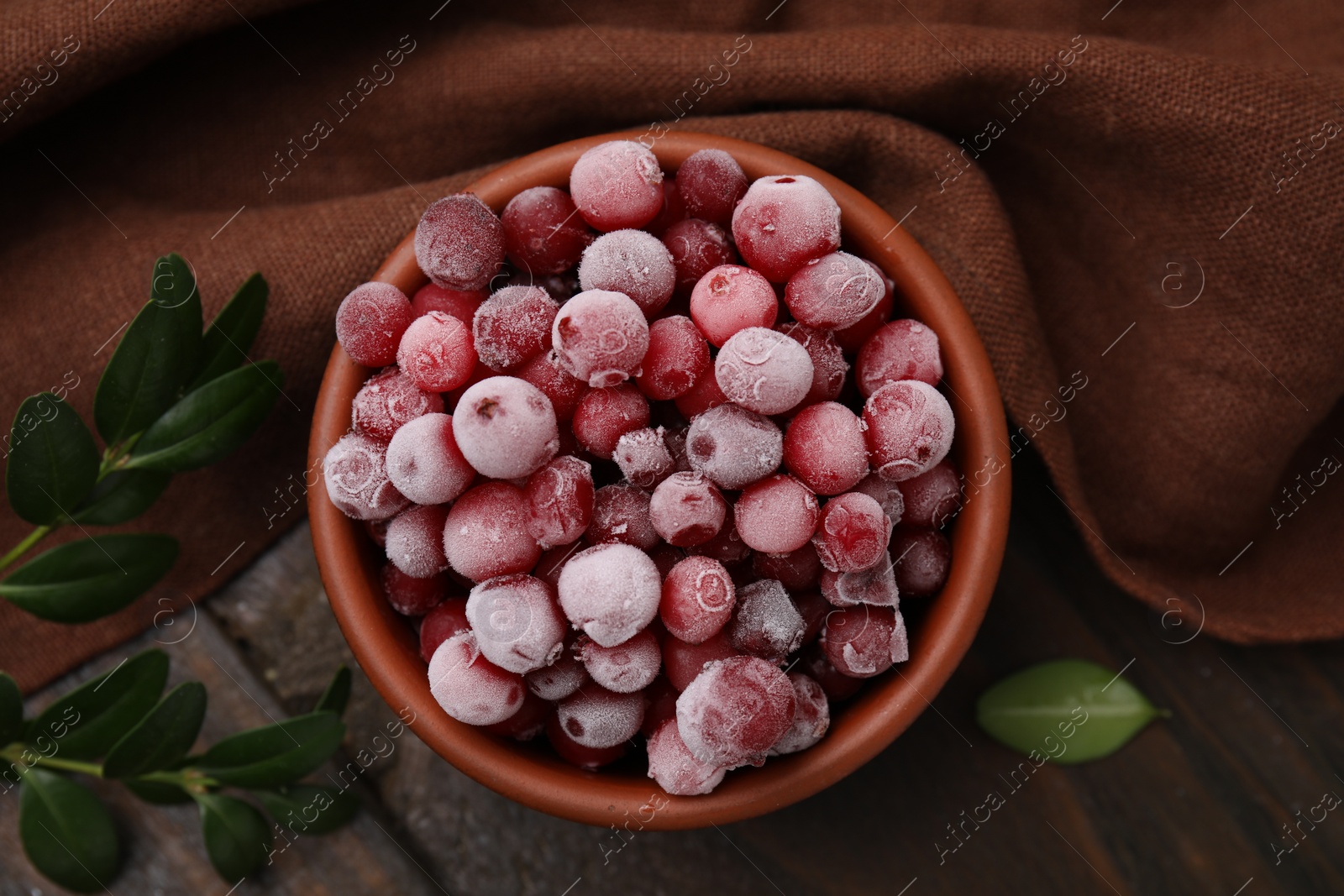 Photo of Frozen red cranberries in bowl and green leaves on wooden table, top view