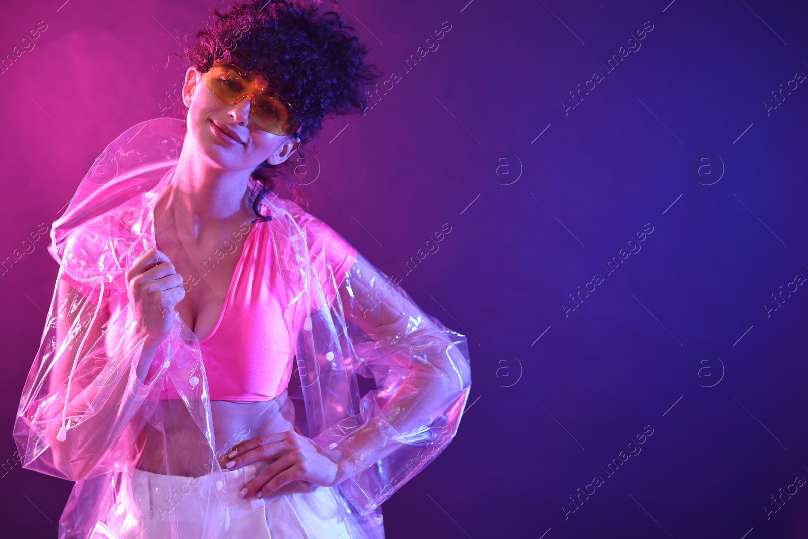 Photo of Beautiful young woman in transparent coat and sunglasses posing on color background in neon lights. Space for text