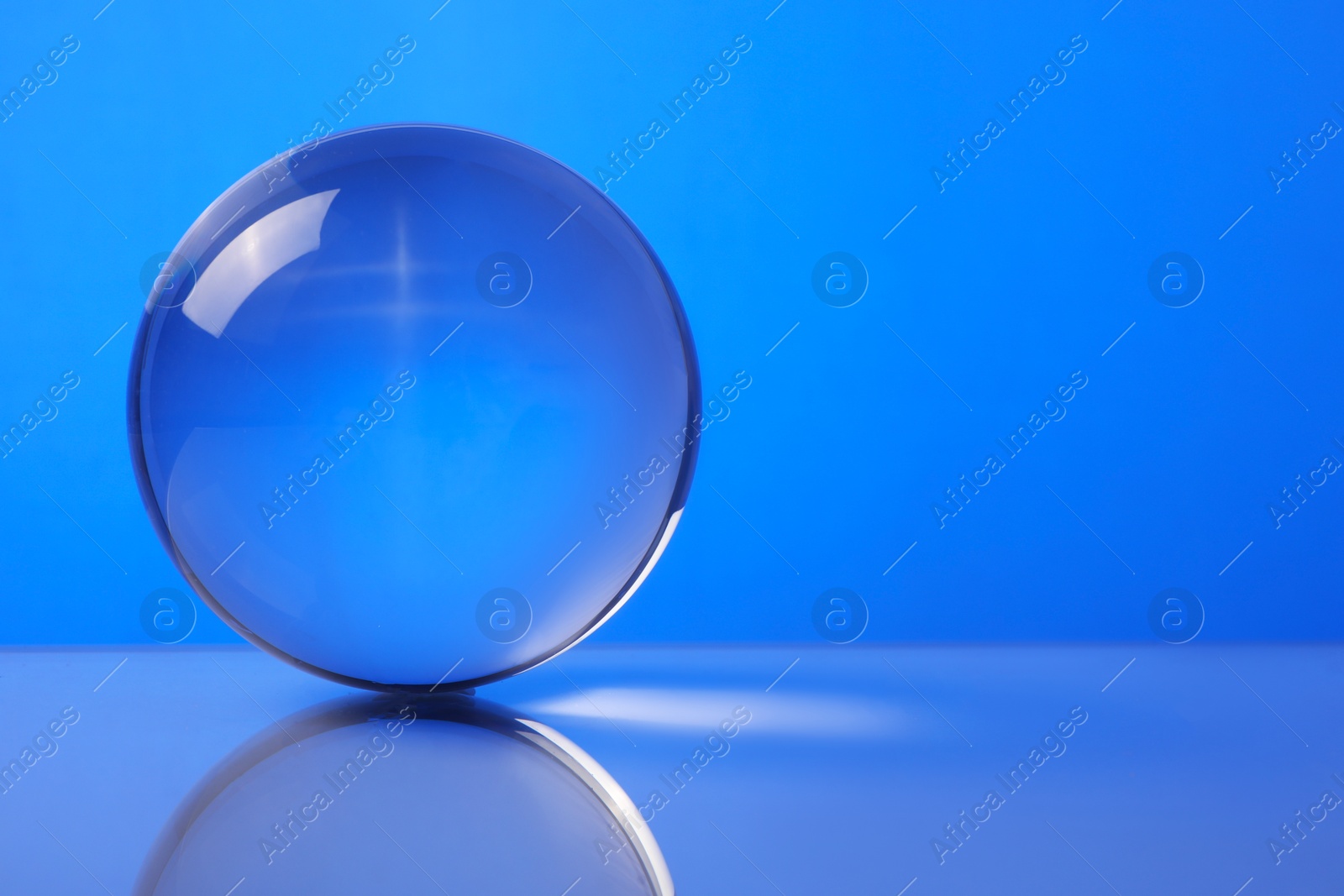 Photo of Transparent glass ball on mirror surface against blue background. Space for text