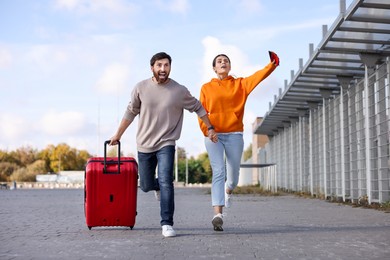 Photo of Being late. Couple with red suitcase running outdoors