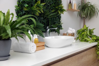 Photo of Green artificial plants, vessel sink and different personal care products in bathroom
