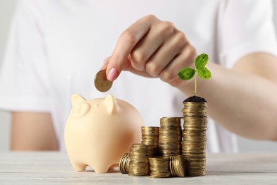 Photo of Woman putting coin into piggy bank near stack with green sprout at white table, closeup. Investment concept