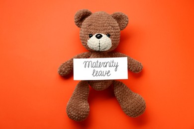 Toy bear and card with phrase Maternity Leave on red background, top view