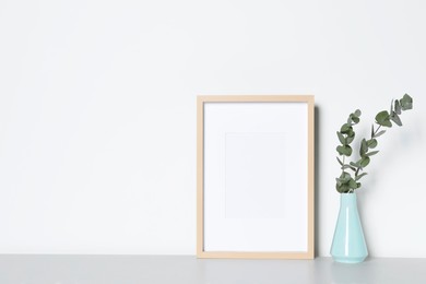 Photo of Empty photo frame and vase with decorative eucalyptus leaves on white table. Mockup for design
