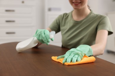 Woman with spray bottle and microfiber cloth cleaning wooden table in room, closeup