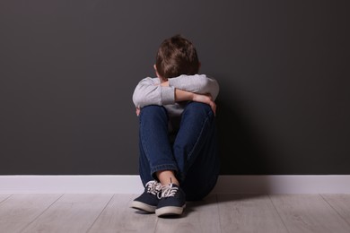 Child abuse. Upset boy sitting on floor near gray wall indoors