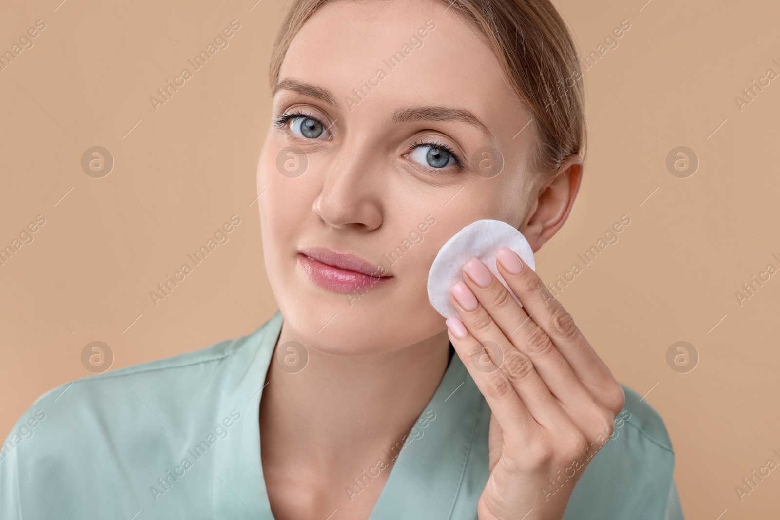 Photo of Young woman cleaning her face with cotton pad on beige background, closeup