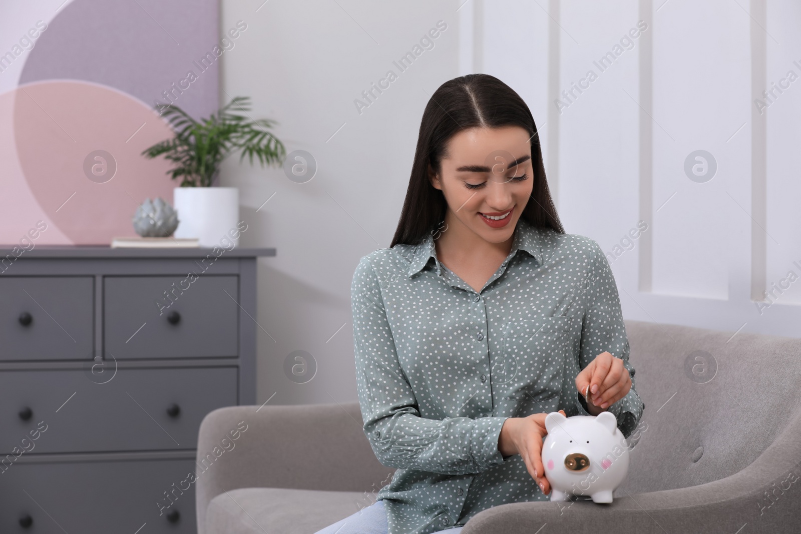 Photo of Young woman putting coin into piggy bank at home