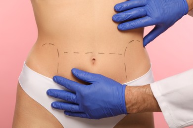 Doctor checking patient's body before cosmetic surgery operation on pink background, closeup