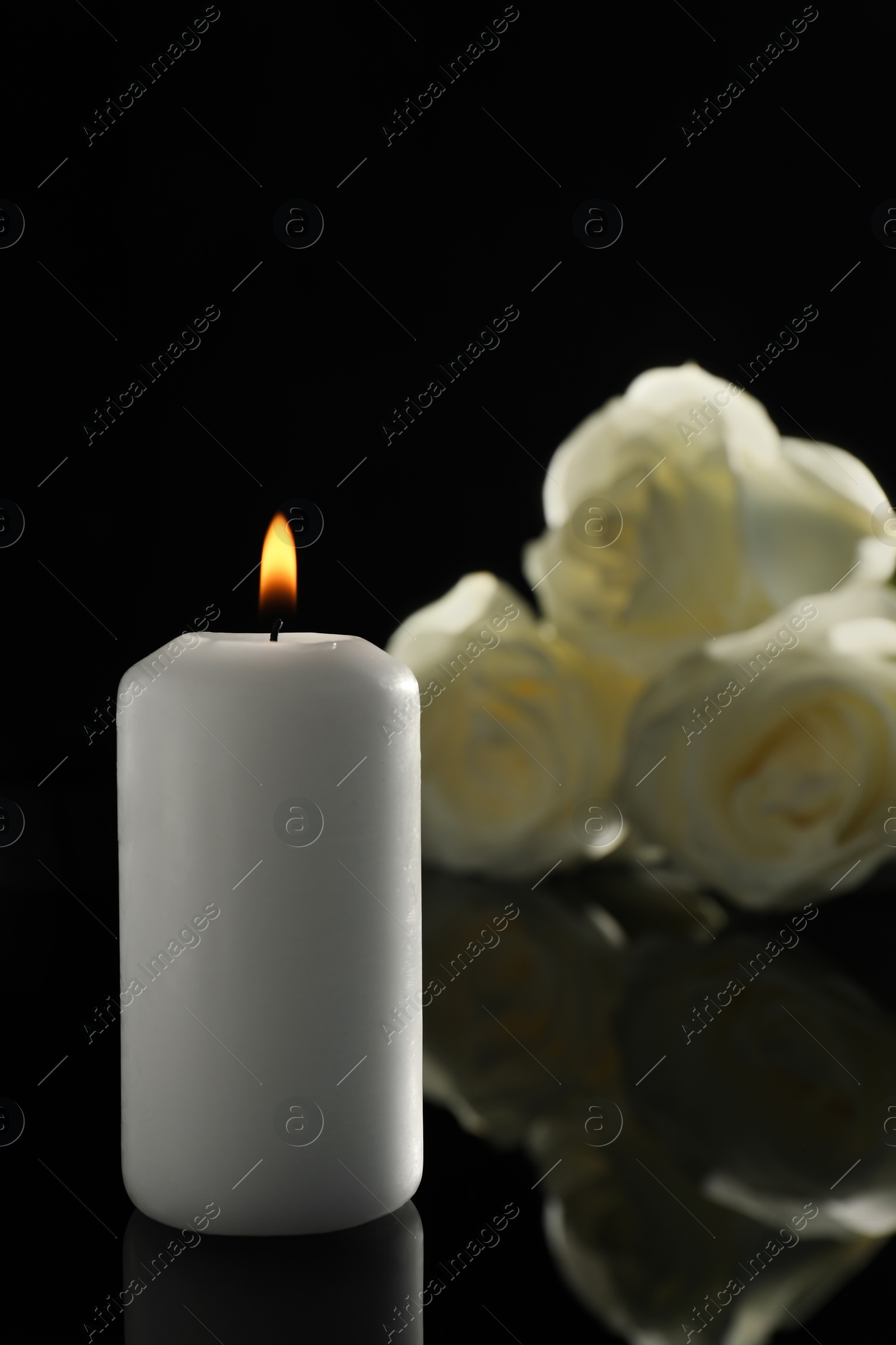 Photo of White roses and burning candle on black mirror surface in darkness. Funeral symbols