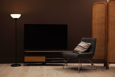 Modern TV on cabinet, armchair and floor lamp near brown wall in room. Interior design