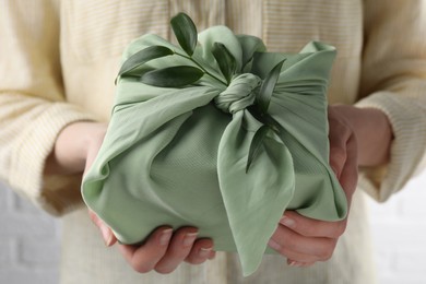 Photo of Furoshiki technique. Woman holding gift packed in green fabric and decorated with ruscus branch, closeup