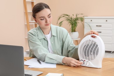 Photo of Young woman adjusting temperature on modern electric fan heater at wooden table indoors