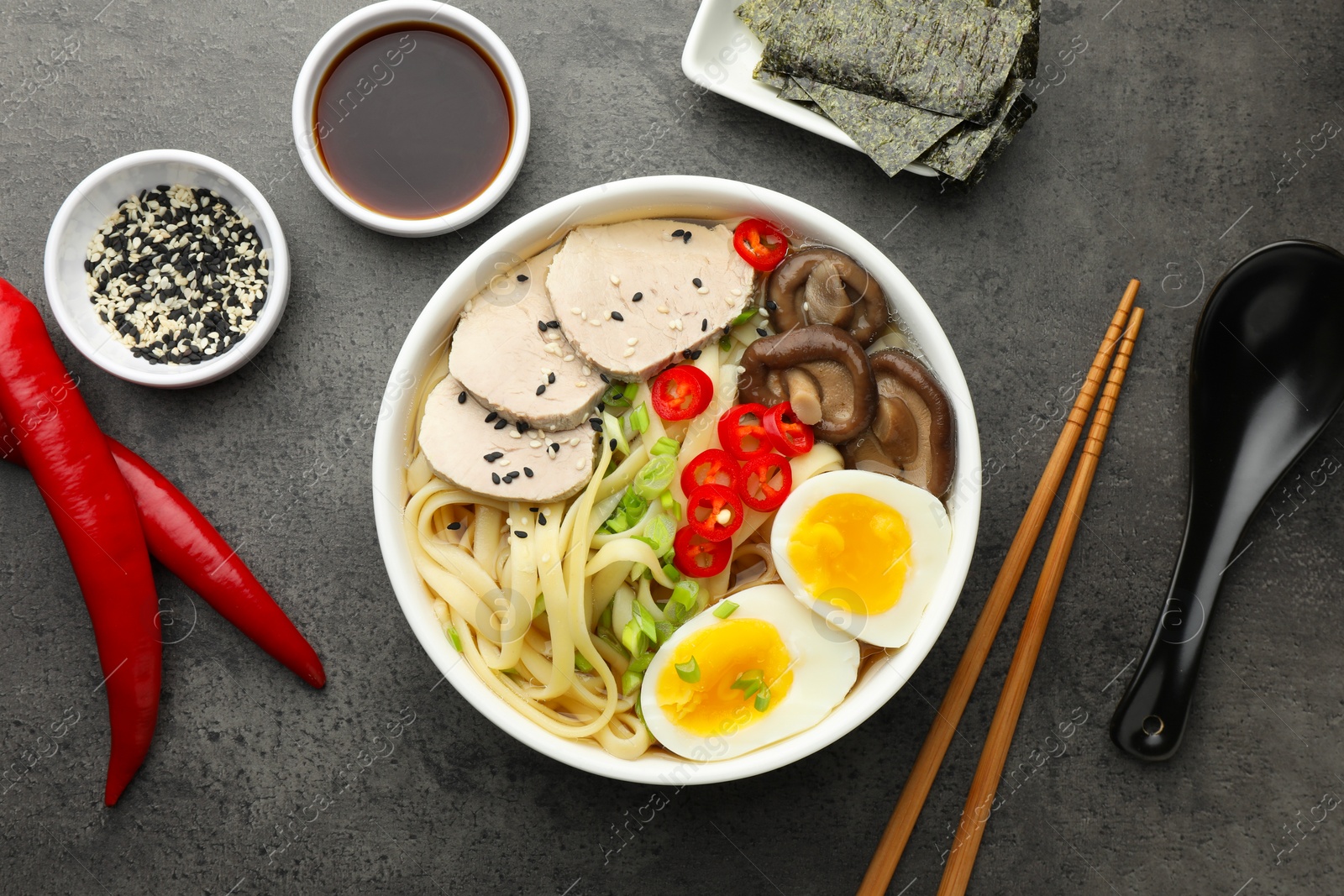 Photo of Delicious ramen with meat in bowl and ingredients on grey textured table, flat lay. Noodle soup