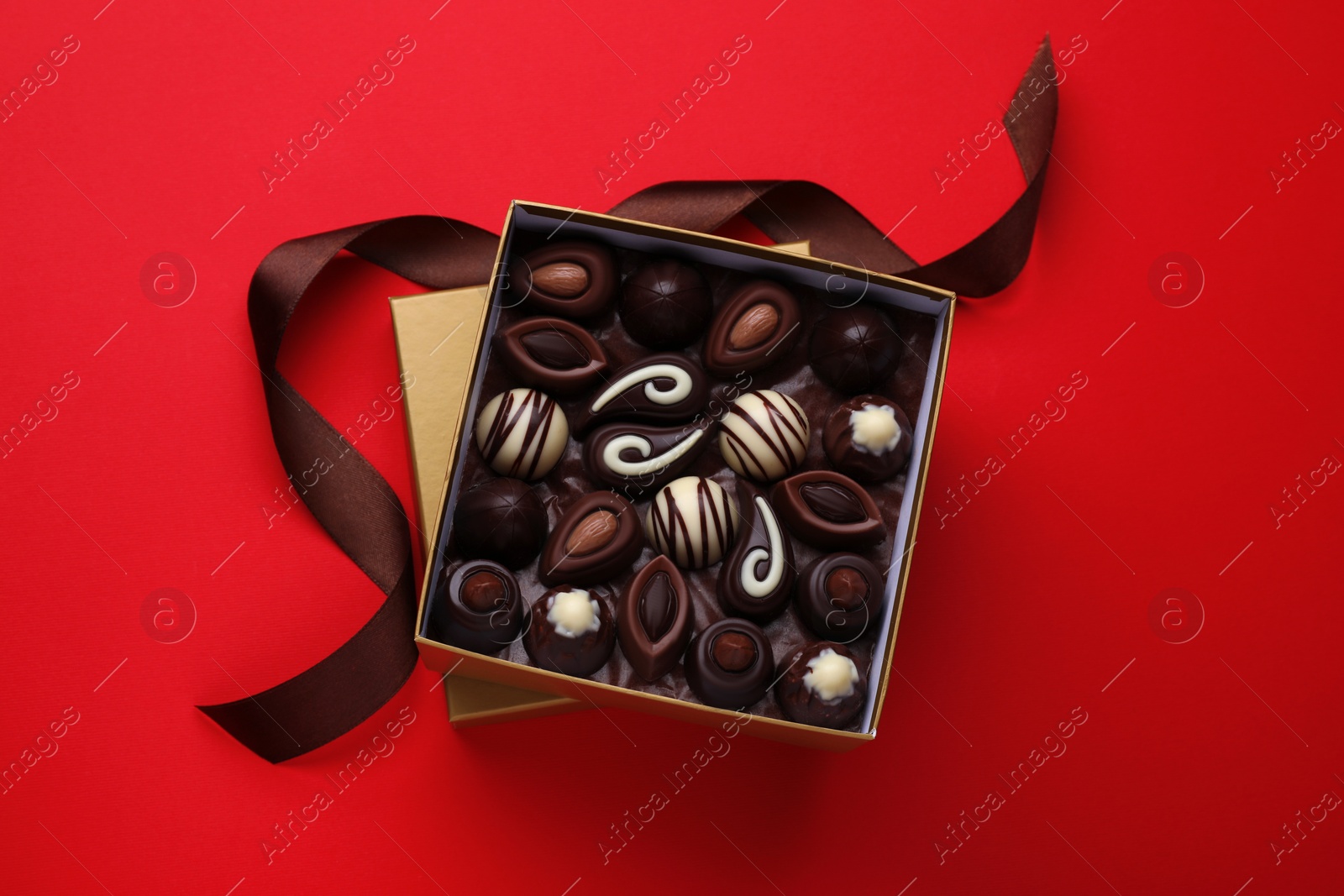 Photo of Box of delicious chocolate candies and brown ribbon on red background, flat lay