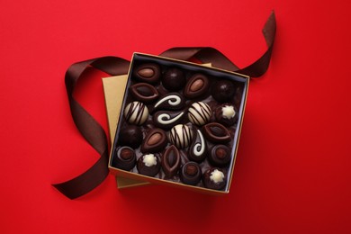 Photo of Box of delicious chocolate candies and brown ribbon on red background, flat lay