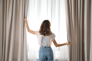 Woman opening window curtains at home in morning, back view