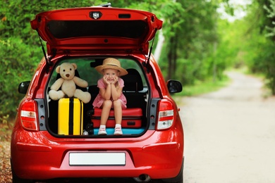 Little girl sitting in car trunk with suitcases near forest. Space for text