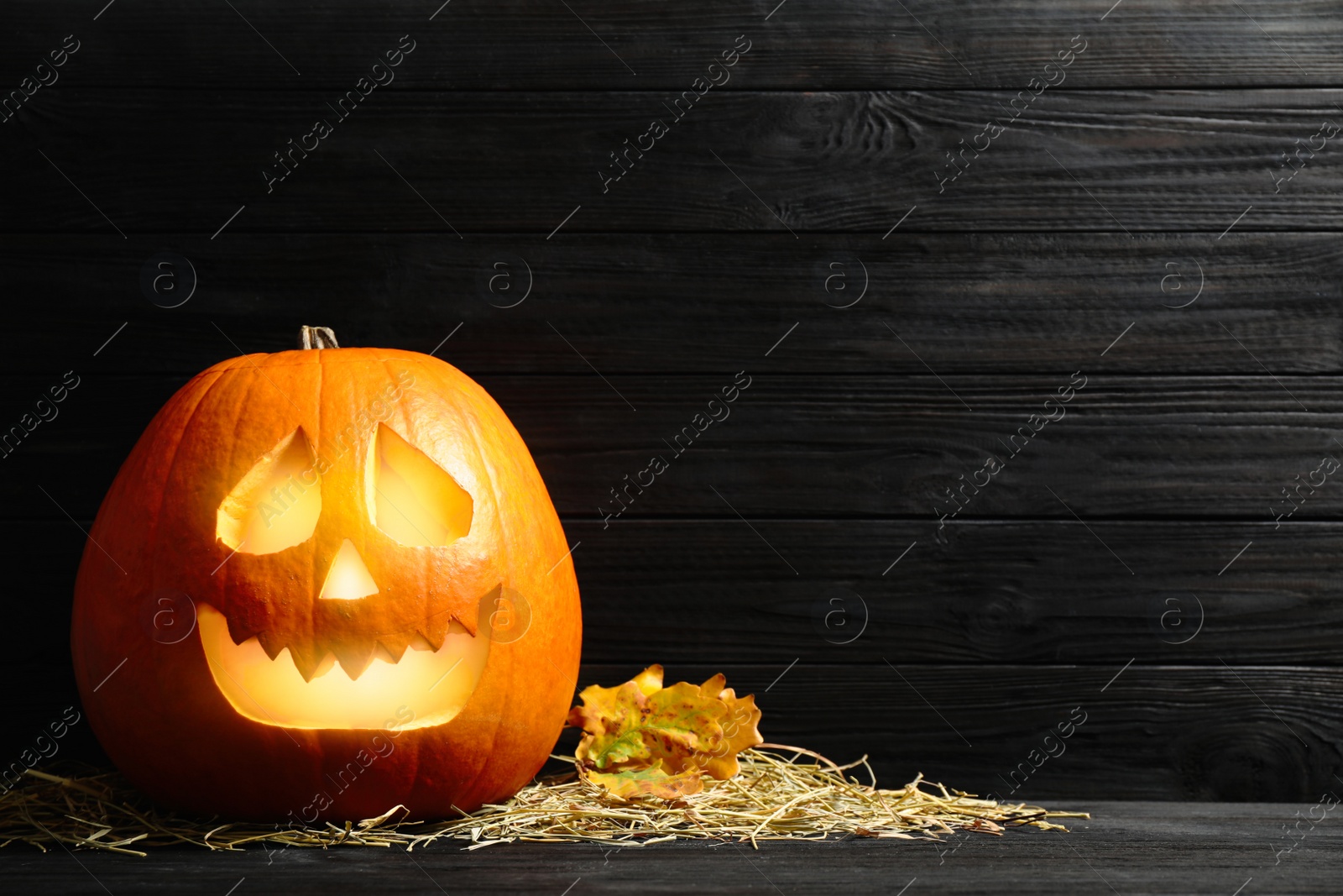 Photo of Pumpkin jack o'lantern, straw and autumn leaves on black wooden table, space for text. Halloween decor