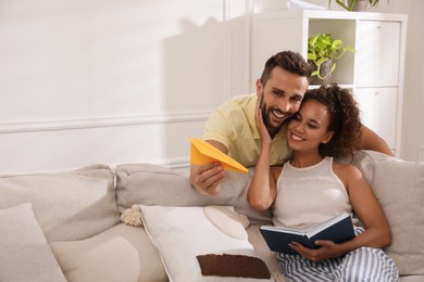 Photo of Happy man playing with paper plane while his girlfriend reading book on sofa in living room
