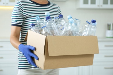 Photo of Woman holding cardboard box with used plastic bottles indoors, closeup. Recycling problem