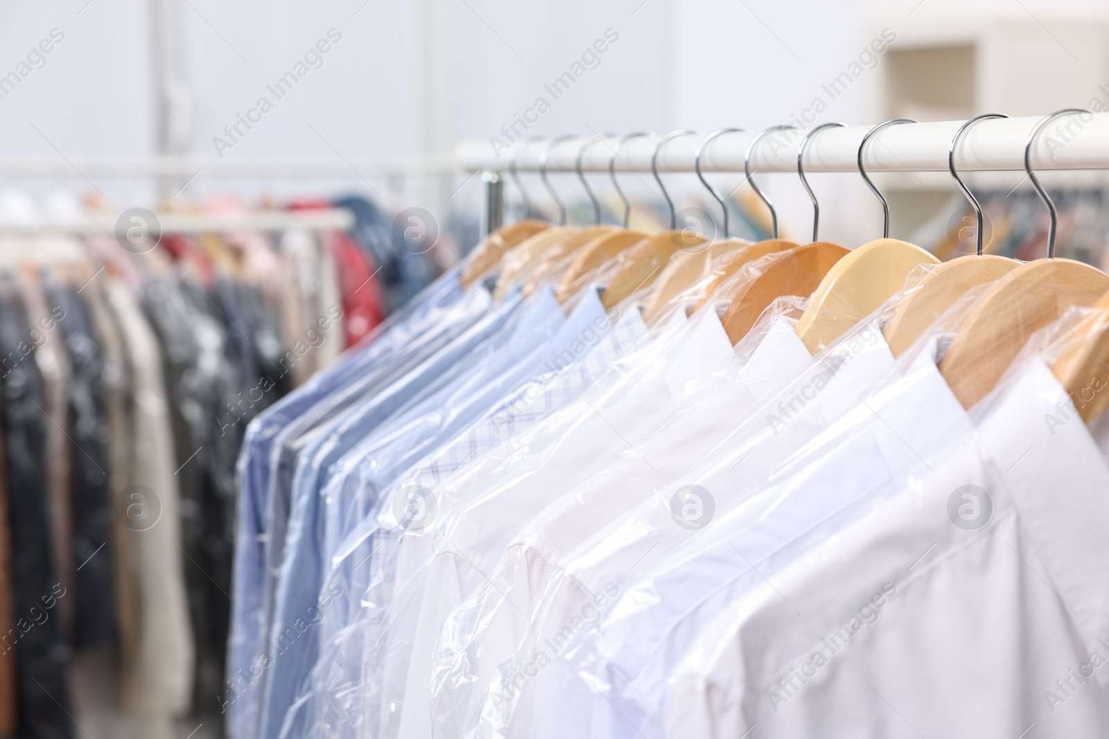 Photo of Dry-cleaning service. Many different clothes in plastic bags hanging on rack indoors, closeup