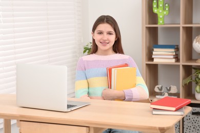 Photo of Cute girl with books near laptop at desk in room. Home workplace