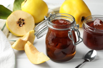 Photo of Tasty homemade quince jam in jars, spoon and fruits on white wooden table, closeup