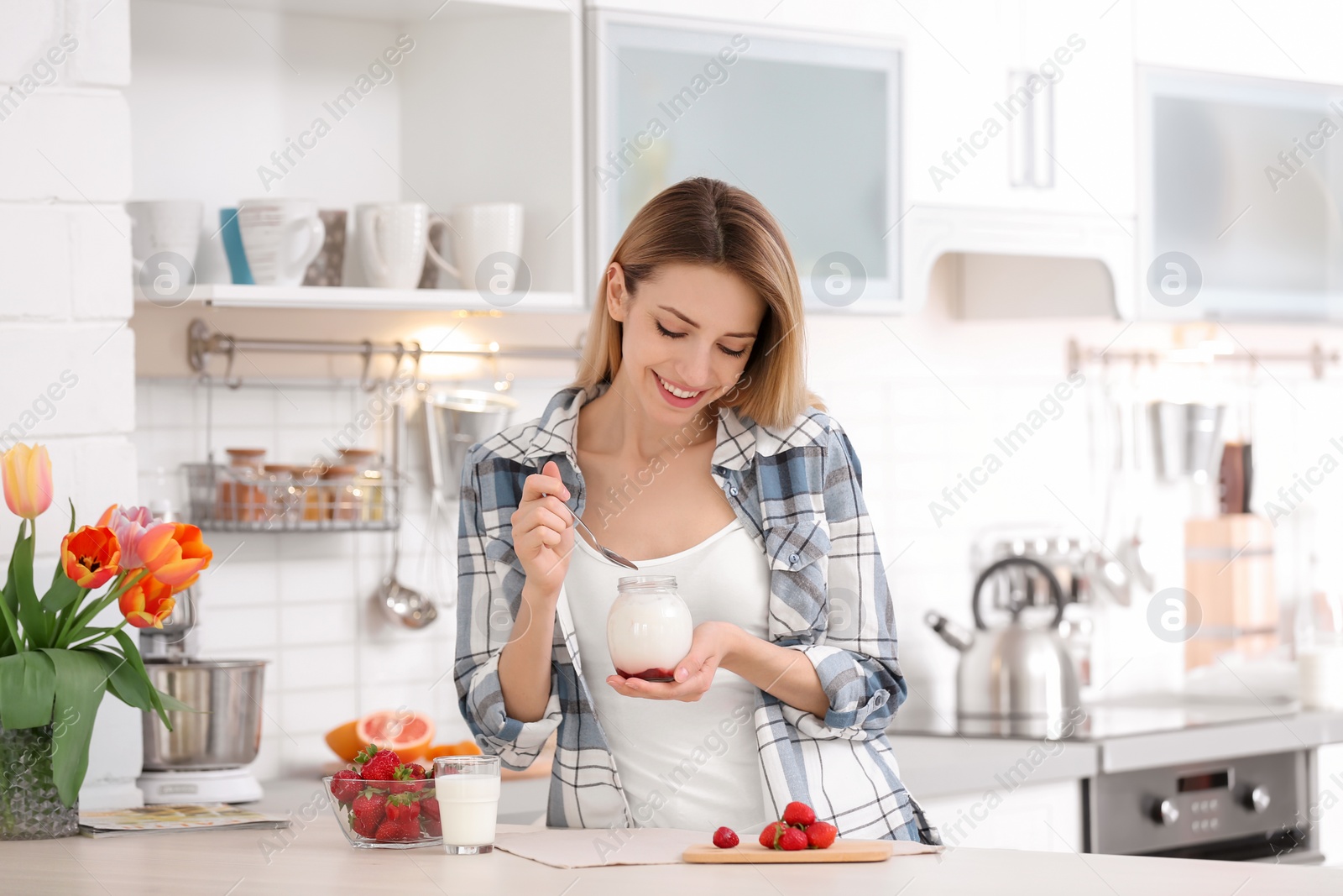 Photo of Young attractive woman eating tasty yogurt at table in kitchen