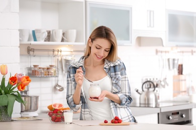 Photo of Young attractive woman eating tasty yogurt at table in kitchen