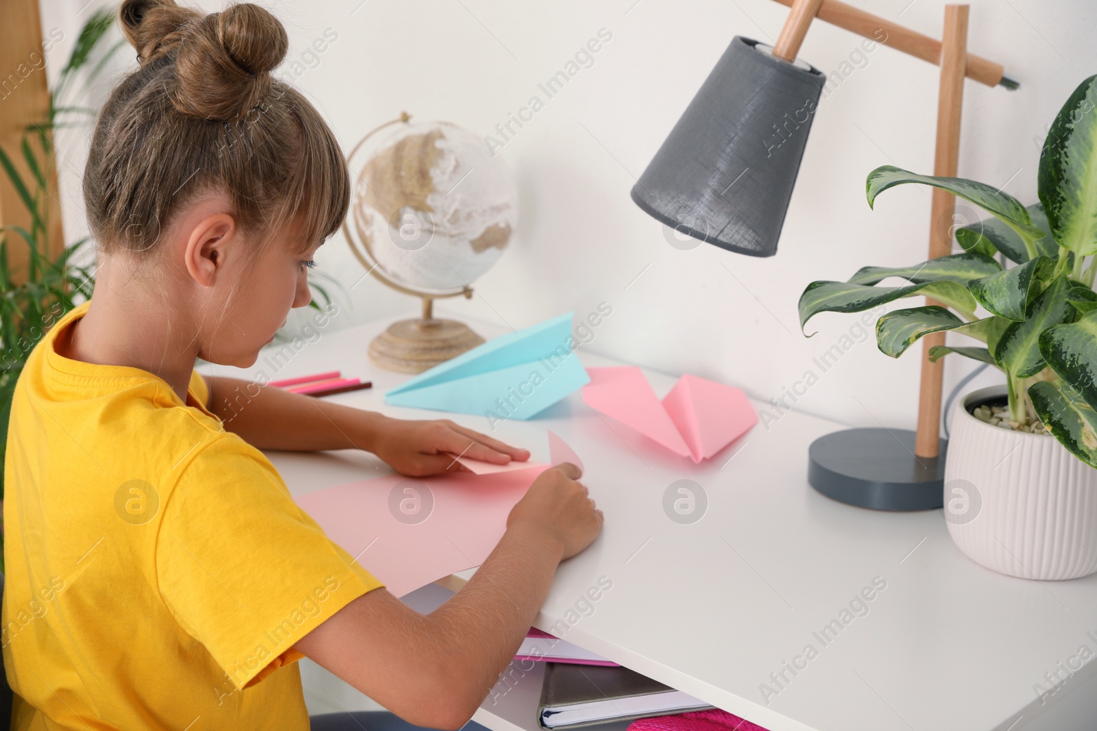 Photo of Cute little girl making paper plane at table in room