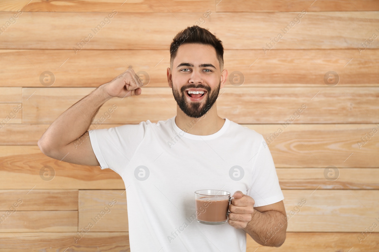 Photo of Young man with glass cup of chocolate milk showing his strength on wooden background