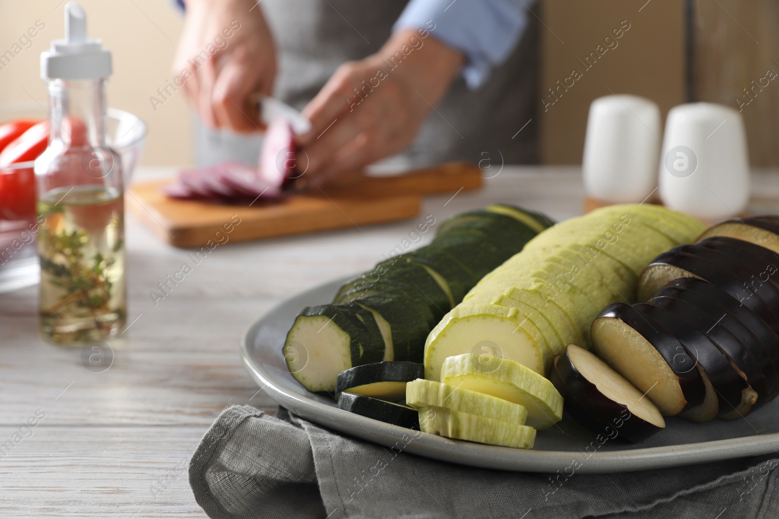 Photo of Cooking delicious ratatouille. Plate with fresh vegetables and woman cutting onion at white wooden table, closeup. Space for text