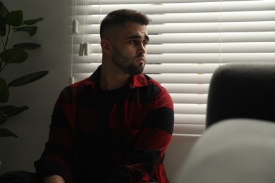 Photo of Sad young man near closed blinds indoors