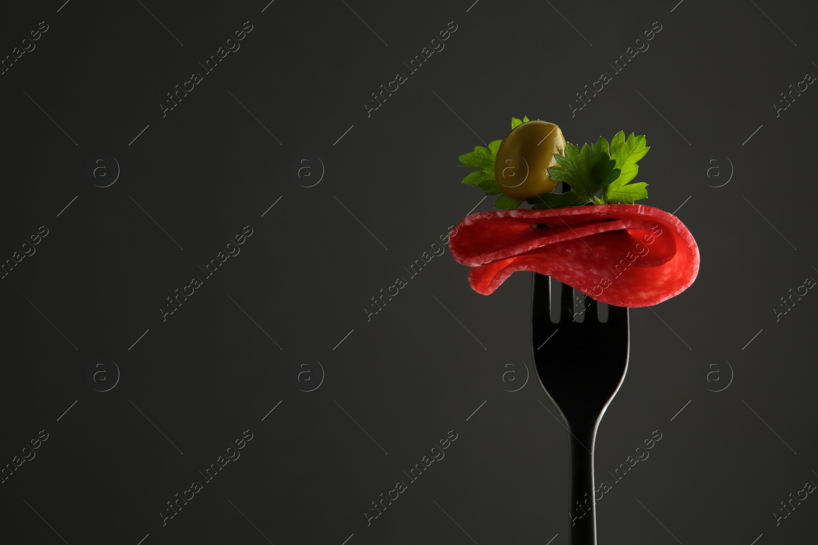 Photo of Fork with tasty slice of salami, olive and parsley on black background, space for text