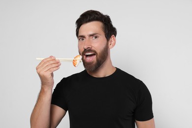 Handsome man eating tasty sushi with chopsticks on light grey background