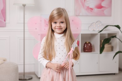Photo of Cute little girl in fairy costume with pink wings and magic wand at home
