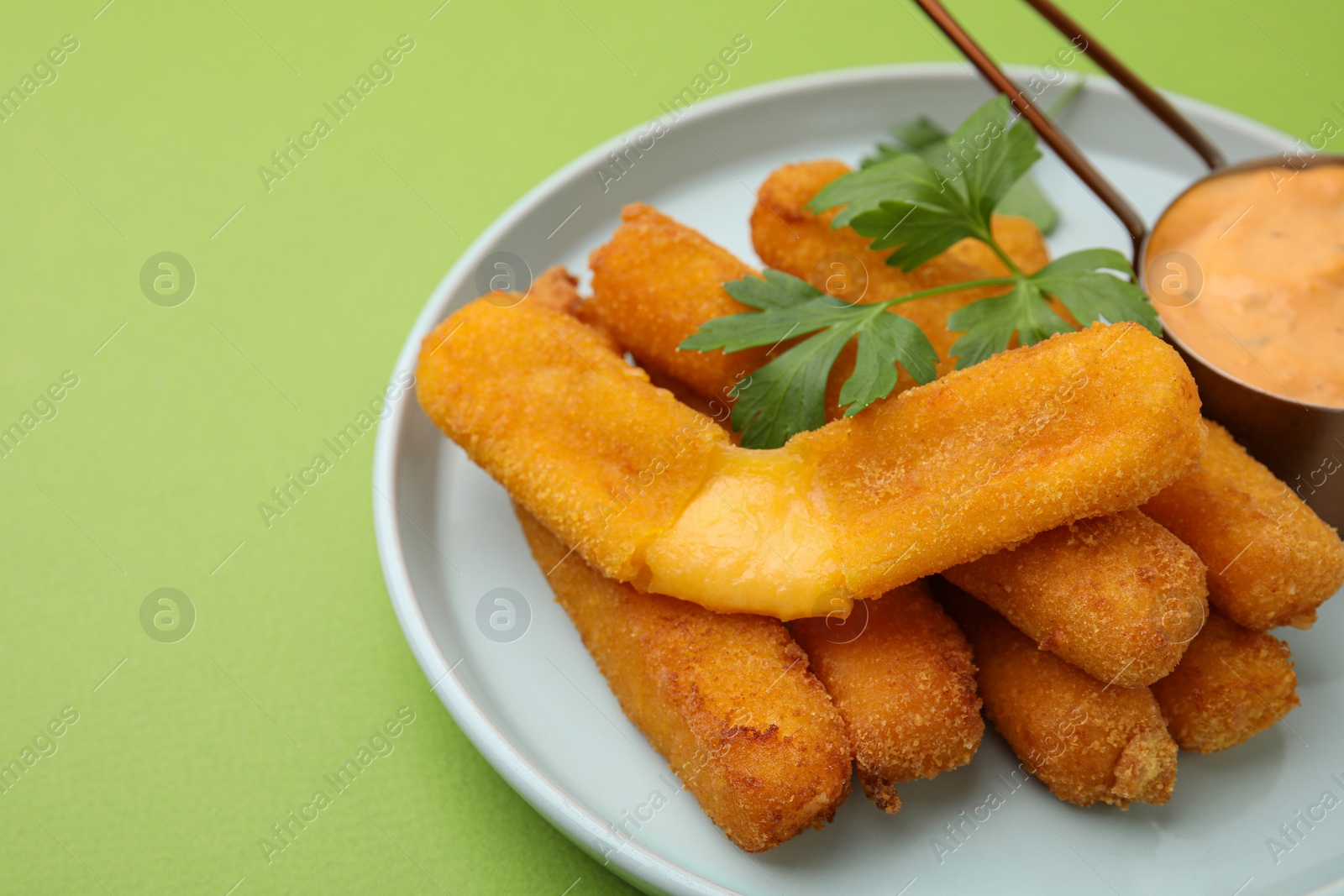 Photo of Tasty fried mozzarella sticks served with sauce and parsley on green background, closeup. Space for text