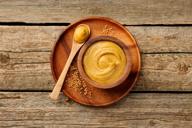 Photo of Spoon and bowl of tasty mustard sauce with dry seeds on wooden table, top view